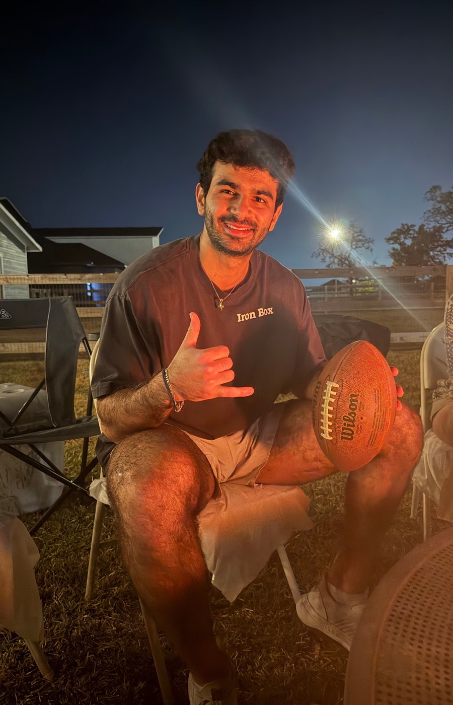 A builder smiling by a backyard fire at night, football in hand